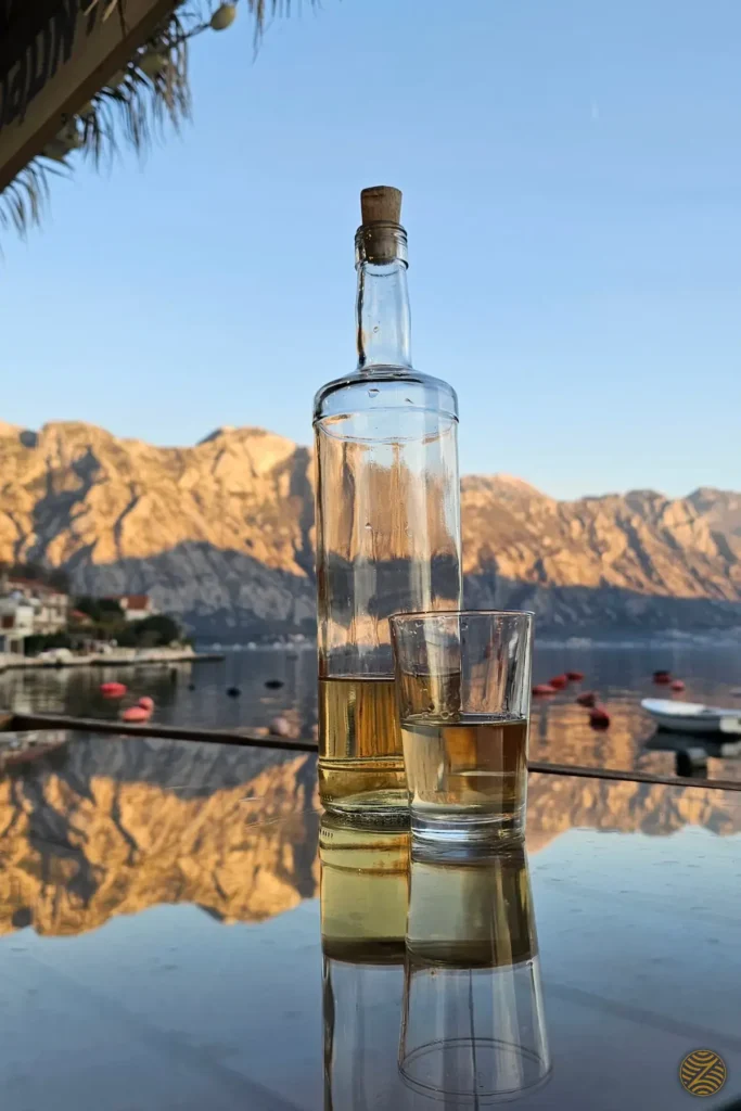 Homemade rakija on a waterfront table reflecting the mountains of the Bay of Kotor