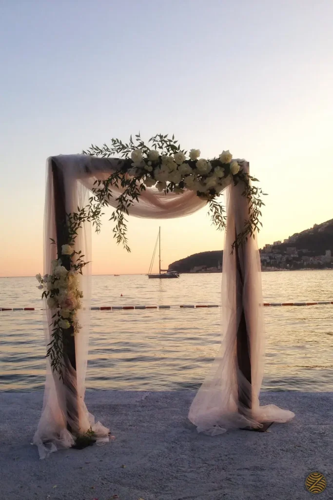 Wedding arch with white roses and tulle at sunset on the seafront, with a sailing yacht and Budva Old Town in the distance